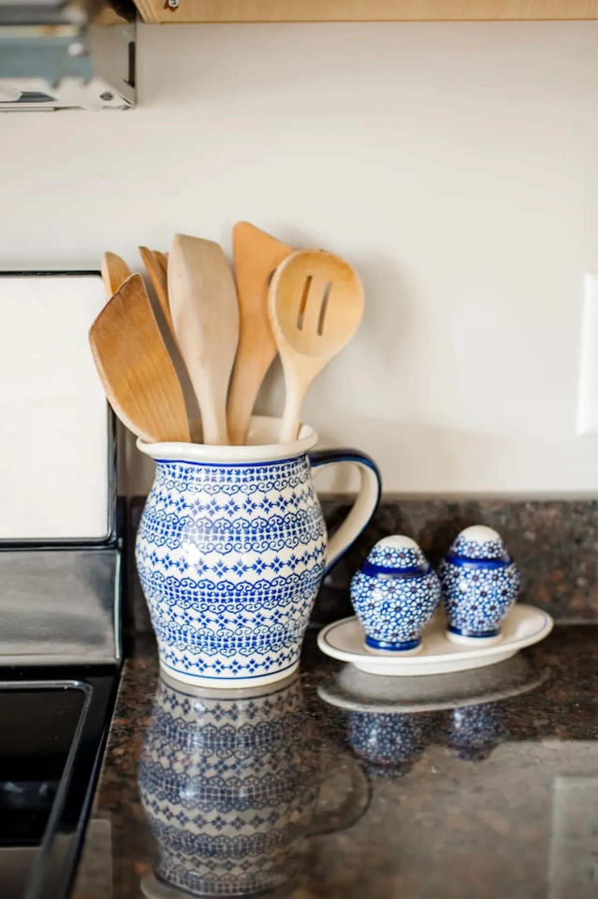 A close-up of a ceramic pitcher with blue patterns, wooden cooking utensils, and decorative salt and pepper shakers arranged on a rustic kitchen counter. The overall setting feels warm and homey, evoking the spirit of heritage recipes and shared meals.