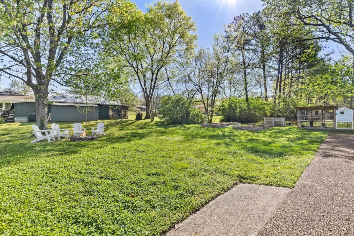 A spacious backyard with lush green grass, adirondack chairs circling a fire pit, trees in the background, and a visible garden area. The scene suggests a variety of activities, from relaxing by the firepit to gardening or playing in the open yard.
