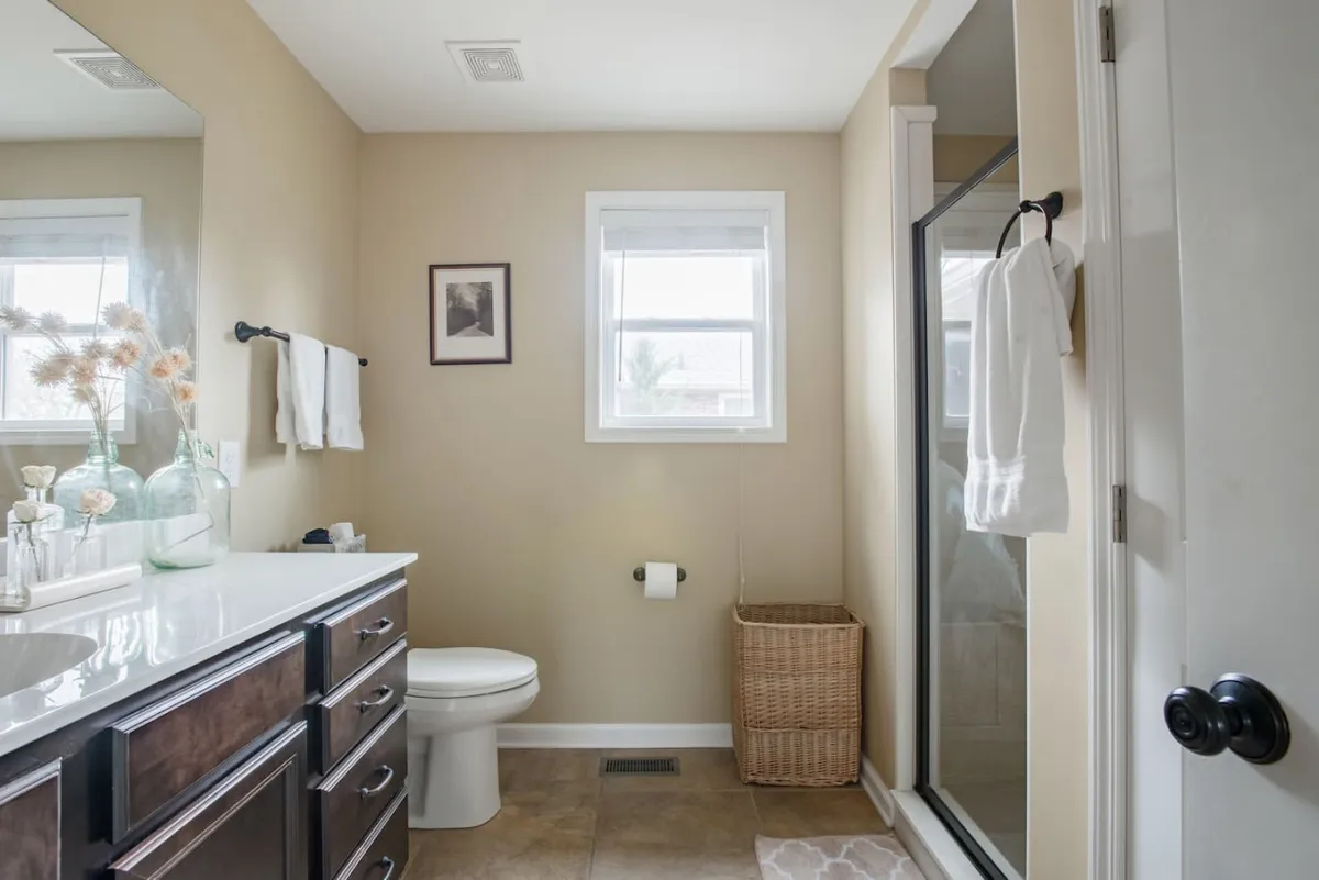 A thoughtfully appointed bathroom featuring a vanity with a large mirror, a shower area, a basket for towels, and a window letting in gentle natural light. The space appears organized, clean, and inviting—ideal for relaxing self-care routines.