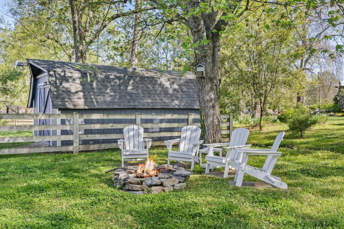 A cozy exterior setup featuring a fire pit surrounded by adirondack chairs under a tree canopy, with a shed in the background. The setting is ideal for stargazing, storytelling, or quiet evenings outdoors.