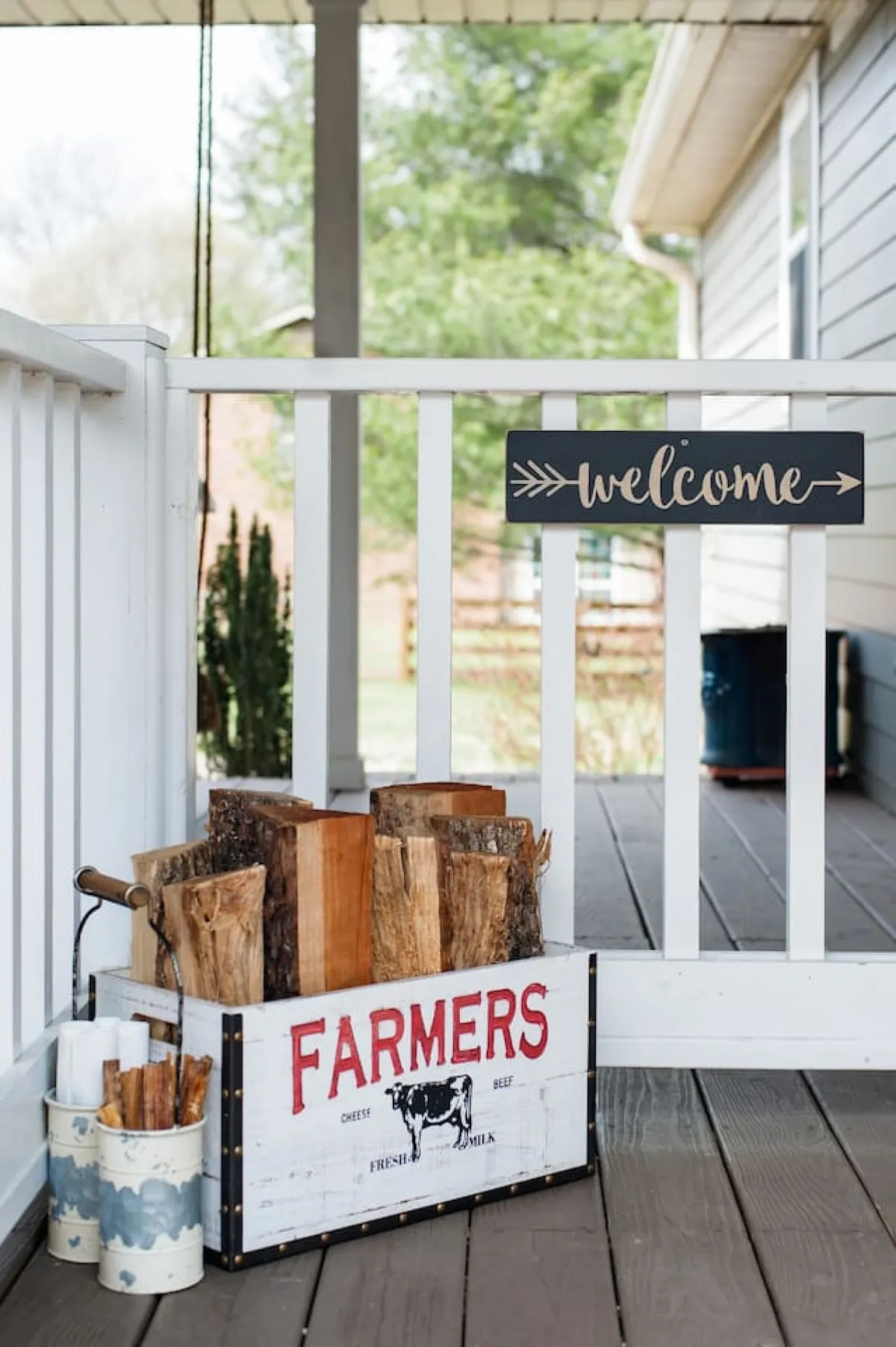 A welcoming outdoor vignette with a rustic welcome sign, wooden log box, and decorative tin containers on a porch. The scene sets a warm, inviting tone for arriving guests.