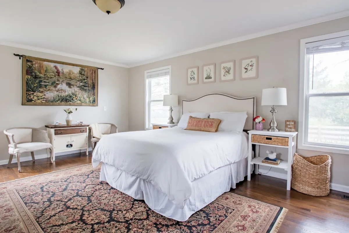 A tranquil bedroom scene featuring a plush bed with layered bedding, vintage artwork on the walls, side tables with lamps, and a patterned rug. Sunlight filters through the window, creating a soft, cozy atmosphere ideal for rest and relaxation.