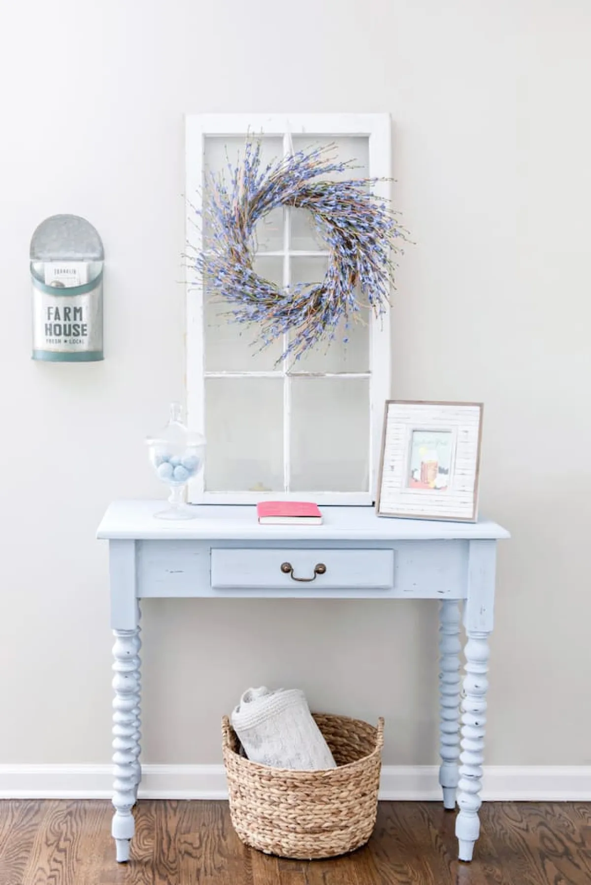 A cozy corner with a blue table adorned with decorative items, a window framed by a wreath, and a welcoming basket with a towel. The setting feels intimate, with soft natural light and a touch of Southern hospitality in the decor.