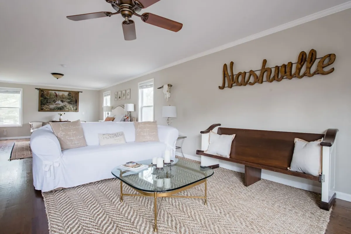 A bright and cozy seating area with a white couch accented by decorative pillows, a glass coffee table, and a visible Nashville sign. The space feels airy and open, ideal for sipping coffee, reading, or socializing with friends and family.