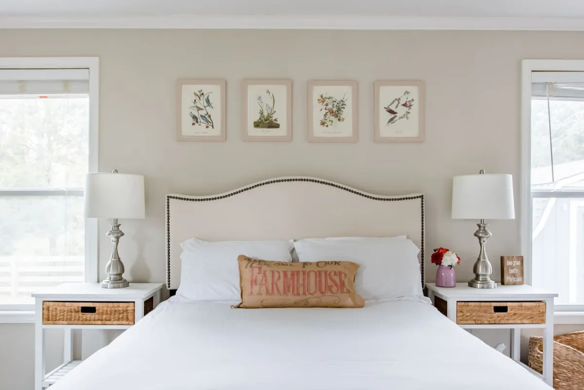 The image showcases a cozy bedroom with a plush bed adorned with decorative pillows, warm table lamps on each side, and curated framed artwork above the headboard. Sunlight streams softly through a nearby window, adding a glow to the inviting space and highlighting the comfortable, farmhouse-inspired decor.