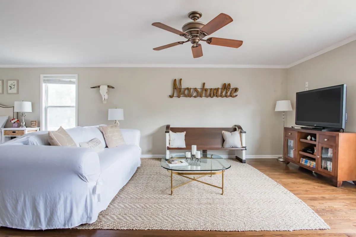 A cozy living room featuring a plush sofa, coffee table, and a prominent 'Nashville' sign on the wall. The TV and decorative accents suggest this is a space for both relaxation and lively gatherings. The arrangement invites conversation and relaxation, perfect for family movie nights or quiet mornings with coffee.