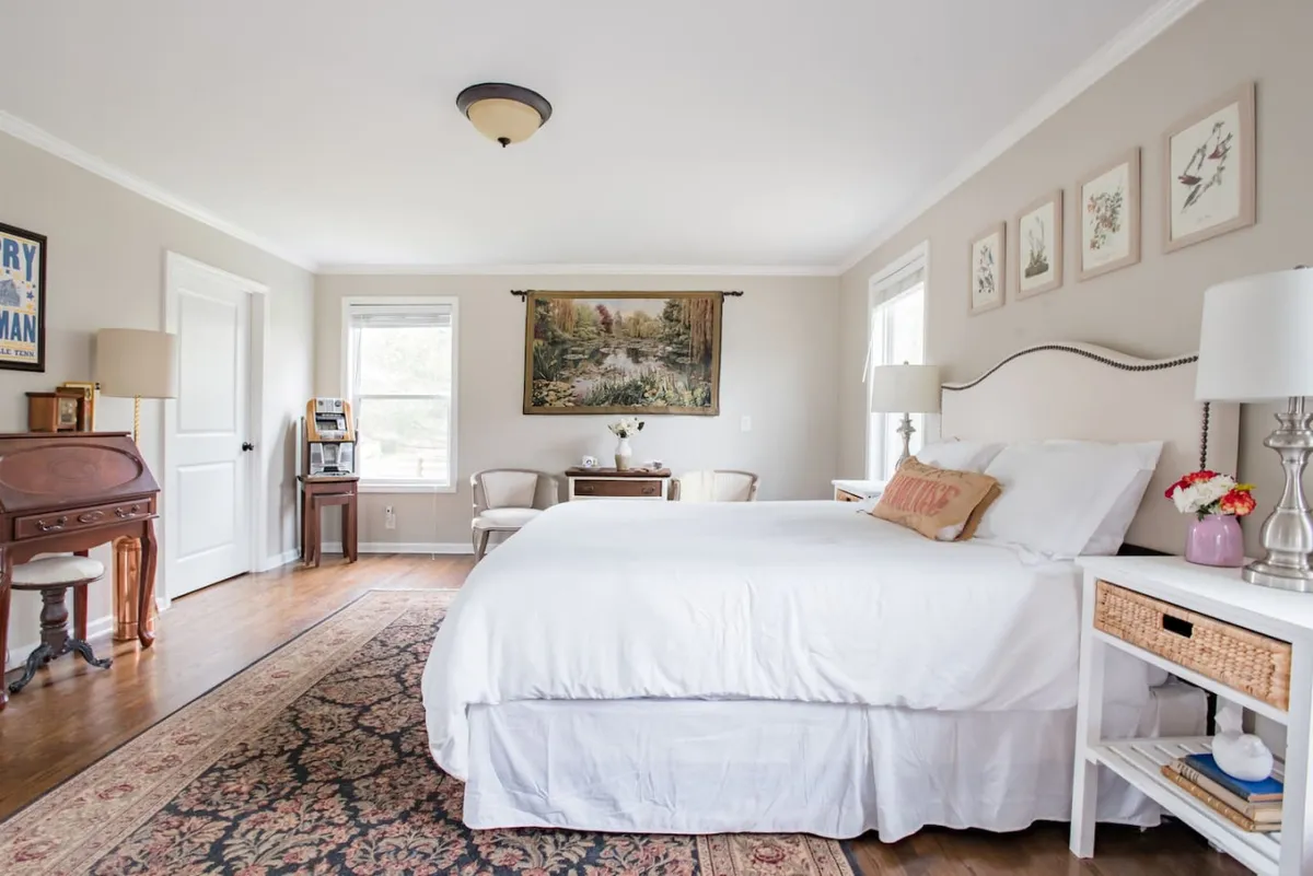 A sunlit bedroom featuring a comfortable bed with fresh linens, a small table with a vase of flowers, and a decorative chair. Soft morning light streams through the window, inviting a leisurely start to the day.