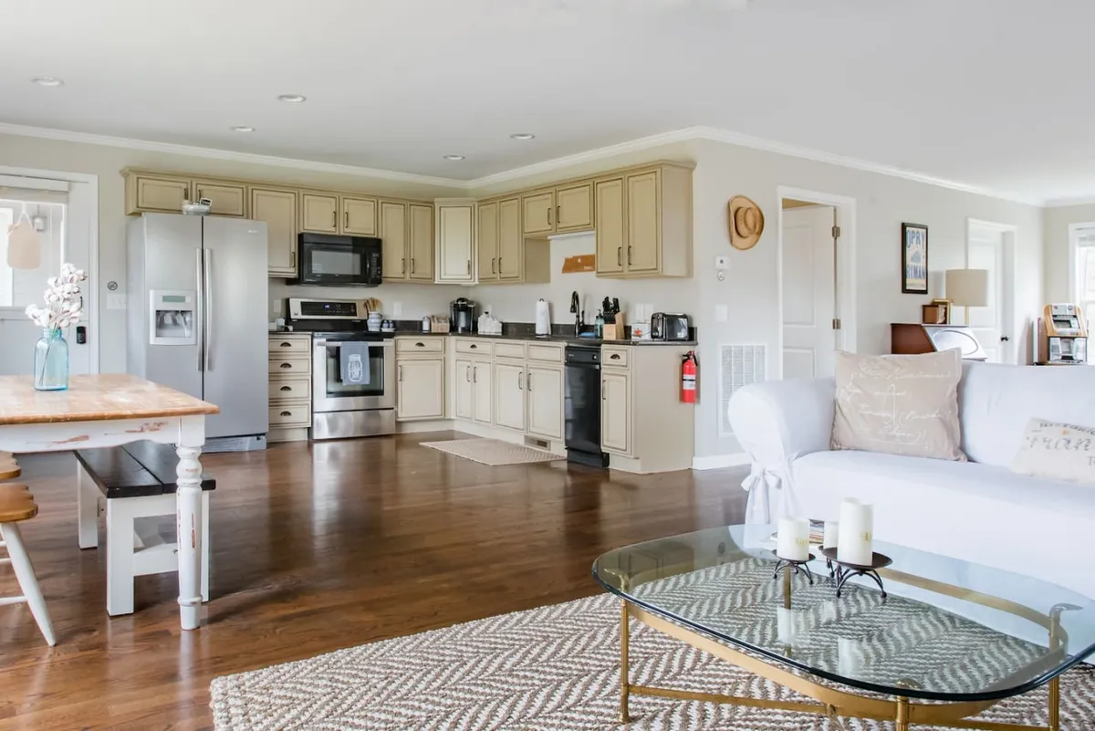 This image captures a welcoming kitchen and living area, complete with kitchen cabinetry, a central island, a cozy sofa, coffee table, and tasteful decorative items. The design suggests a space for socializing and sharing meals, echoing a farmhouse's communal heart.