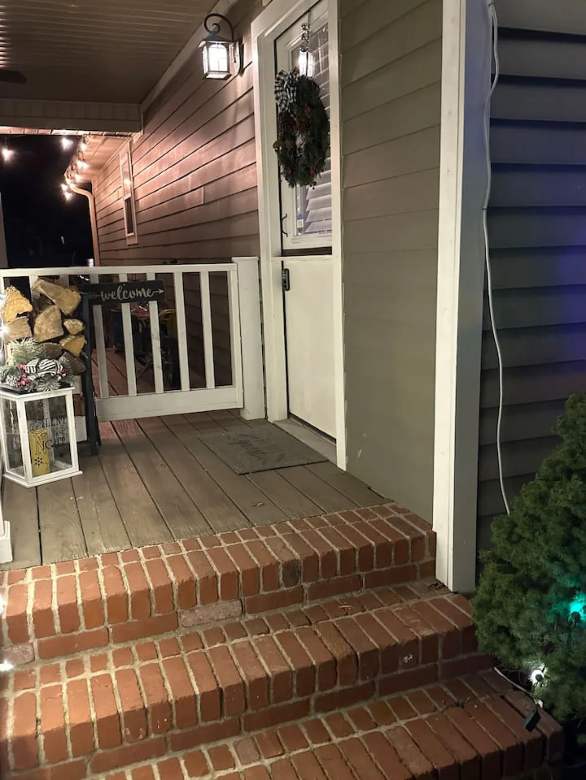 The image presents an inviting front door with a festive wreath, a welcoming sign, and lanterns on brick steps leading into the home. The entryway exudes warmth and hospitality, suggesting a friendly farmstead atmosphere.