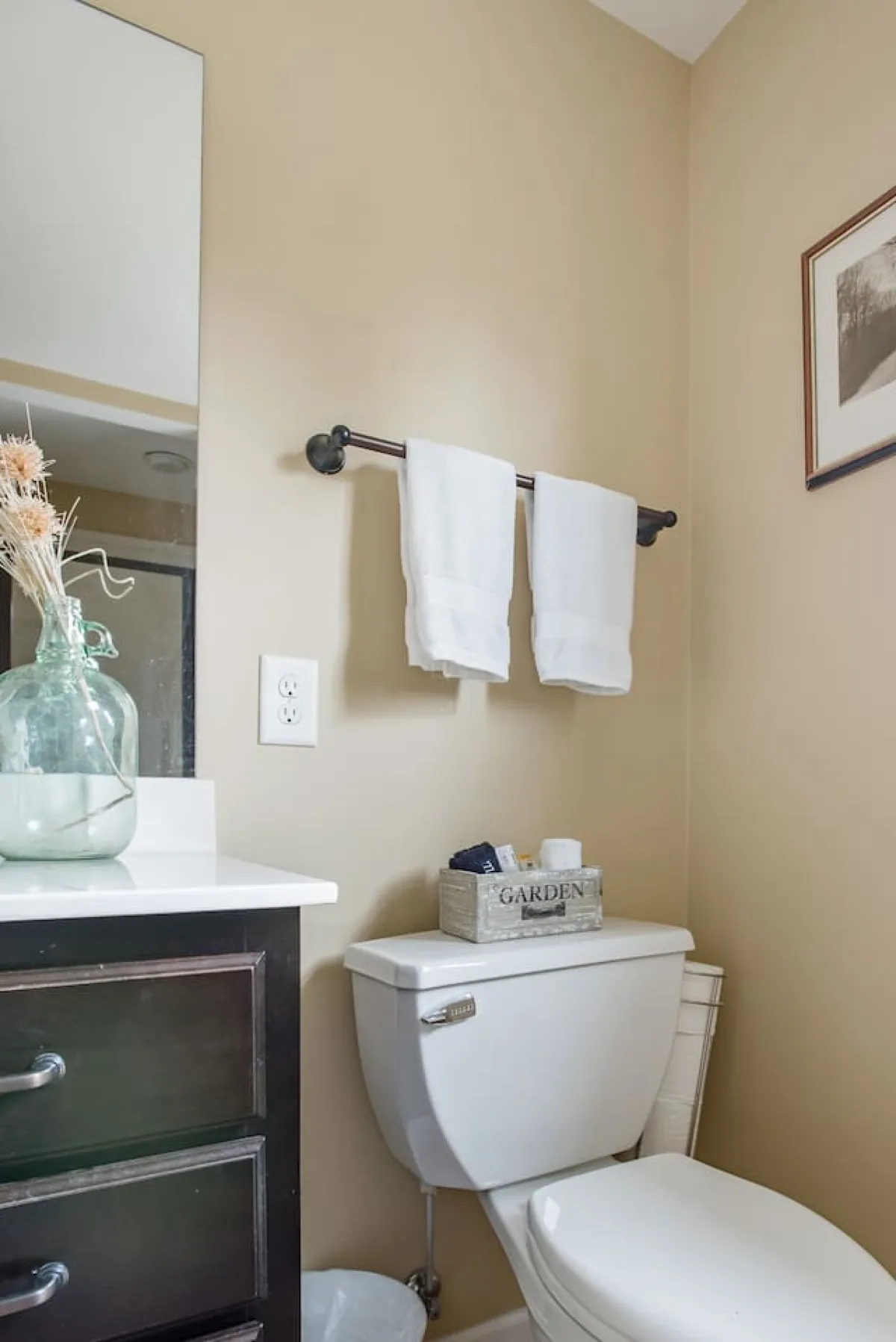 A serene bathroom scene with a glass vase of dried flowers, neatly folded white towels, and a garden-themed storage box, all bathed in soft natural light. The mood is tranquil and encourages relaxation and self-care.
